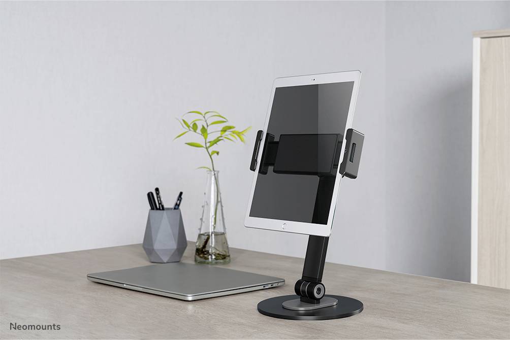 A tablet is positioned on an adjustable desk stand next to a laptop in a modern office, with a vase containing a plant in the background.