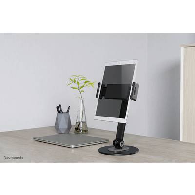 A tablet is positioned on an adjustable desk stand next to a laptop in a modern office, with a vase containing a plant in the background.