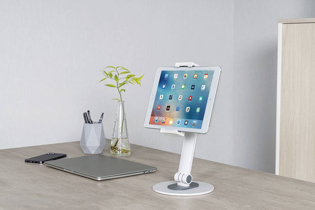 On a desk stands a tablet in a white stand next to a laptop, a mobile phone and a vase with branches.
