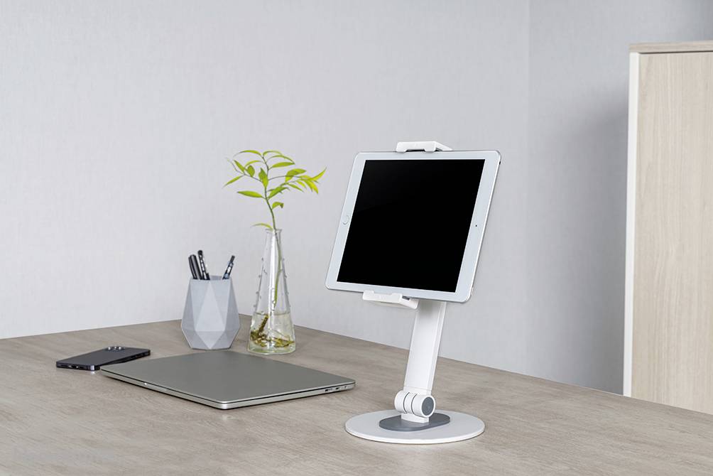 A desk with a tablet on a stand, a laptop, a smartphone, and a pen holder with a plant decoration.