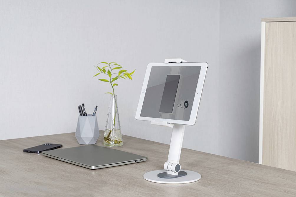A tablet is sitting on a white stand on a desk. Next to it are a laptop and a smartphone. In the background, there is a vase with a plant.