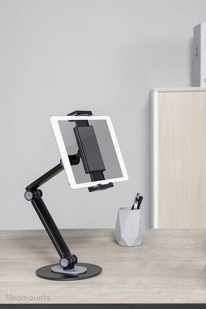 A tablet is mounted in an adjustable holder on a desk. A pencil pot stands in the background.