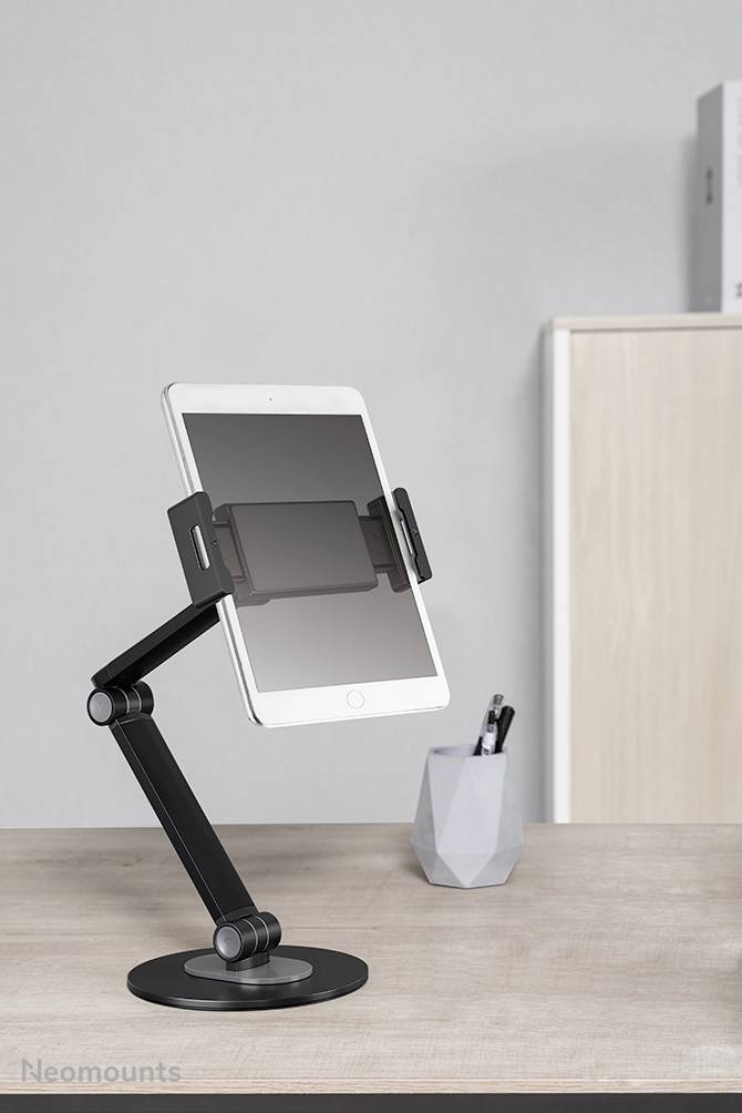 'A tablet is positioned in an adjustable black holder with an arm on a desk. In the background are a pen holder and a cabinet.'
