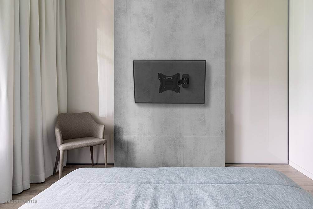 A modern bedroom with a grey wall panel and a television mount. On the left is a beige armchair, and on the right is a large window.