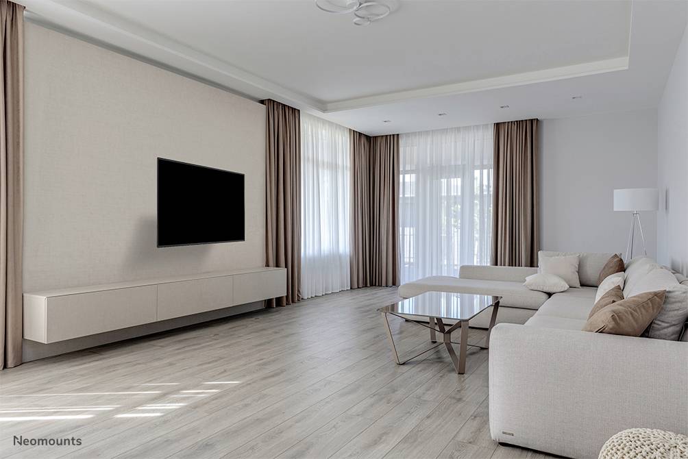 Bright living room with a modern sofa, coffee table and large wall-mounted television, framed by floor-length curtains in front of expansive windows.