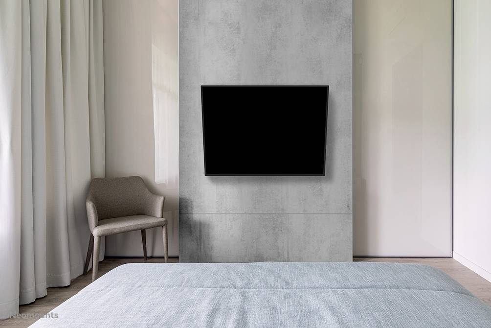 A modern bedroom with a grey wall, on which a large flat-screen television is mounted. A chair stands in the corner next to light curtains.