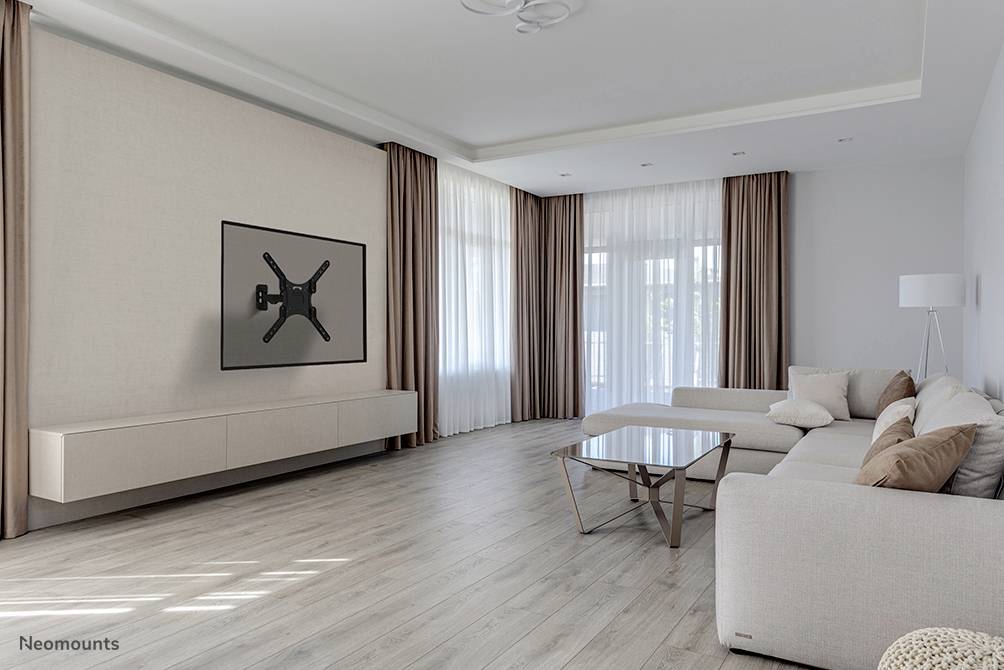 A modern living room with a grey sofa, a glass coffee table and a large television mounted on the wall. Large windows with long curtains.