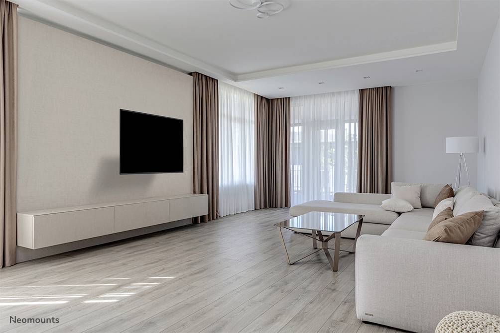 Bright living room with a large corner sofa, glass coffee table and wall-mounted TV. Large windows with floor-length curtains allow plenty of light to enter.