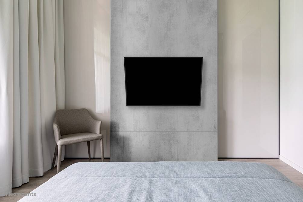 A modern bedroom with a grey concrete accent wall, on which a large, black flatscreen television is mounted. A chair stands to the left.