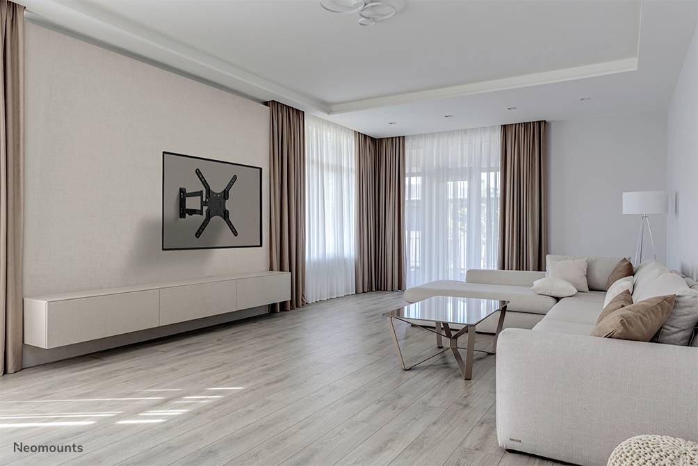 Living room with light-coloured walls and wooden flooring. A large sofa with cushions stands in front of a wall-mounted TV bracket without a television. Large windows with long curtains allow light to flood in.