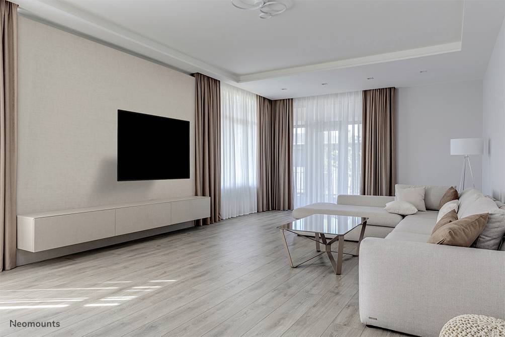 Spacious living room with light wooden flooring, beige sofa with cushions, glass coffee table, wall-mounted television and large windows.