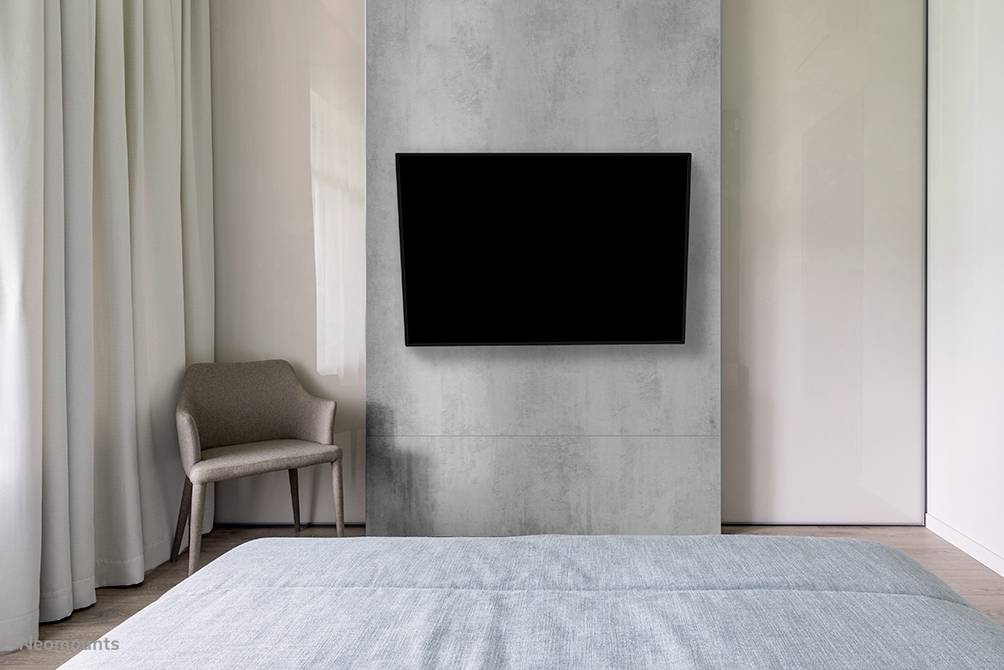 A modern bedroom with a television mounted on the wall, a chair and a bed in the foreground. Neutral colours dominate the room.