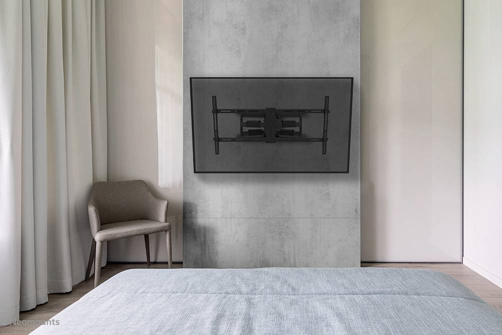 A view into a modern bedroom with a grey concrete wall, on which a flat-screen television is mounted. A chair stands in the corner next to curtains.