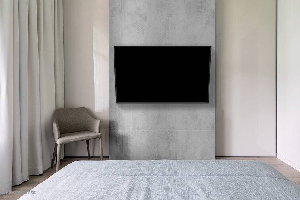 Simple bedroom with a grey wall, on which a flat-screen television is mounted. On the left, an armchair stands next to long curtains.