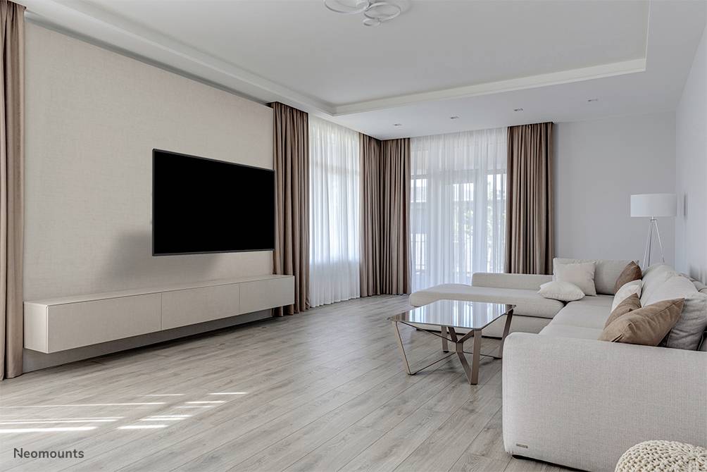 Bright living room with a large sofa, coffee table, and wall-mounted television. Large windows with curtains and wooden flooring.