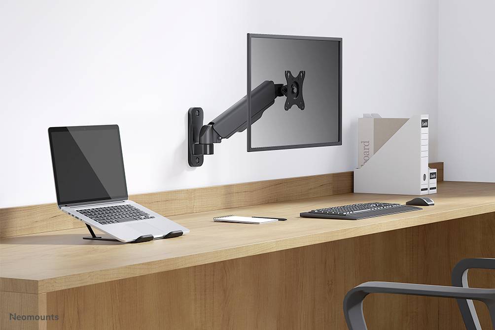 A desk with a laptop standing upright, a keyboard, and a monitor mounted on an adjustable wall bracket.