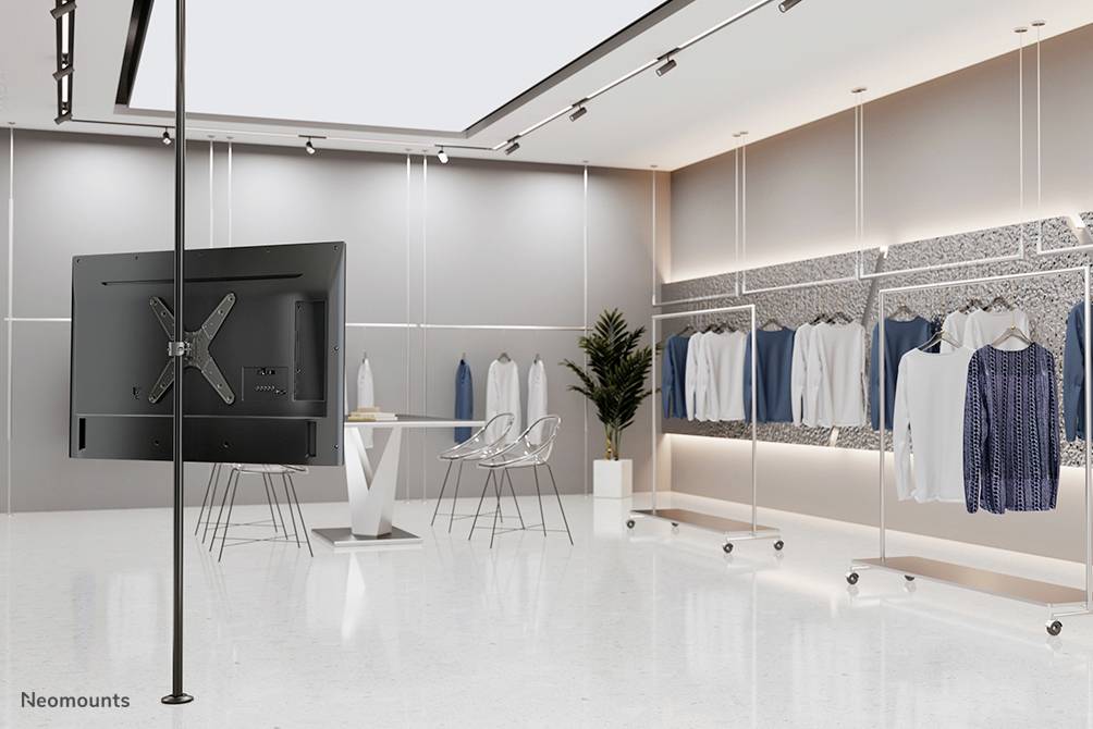 A contemporary showroom with hanging garments, a table and chairs. A screen mounted on the ceiling. Decorative plants on the right.