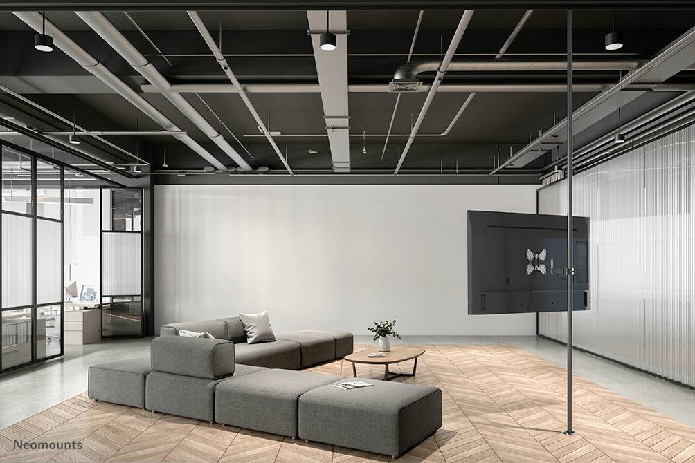 A modern office space with grey modular sofas on a wooden floor. A large screen is mounted on the right wall.