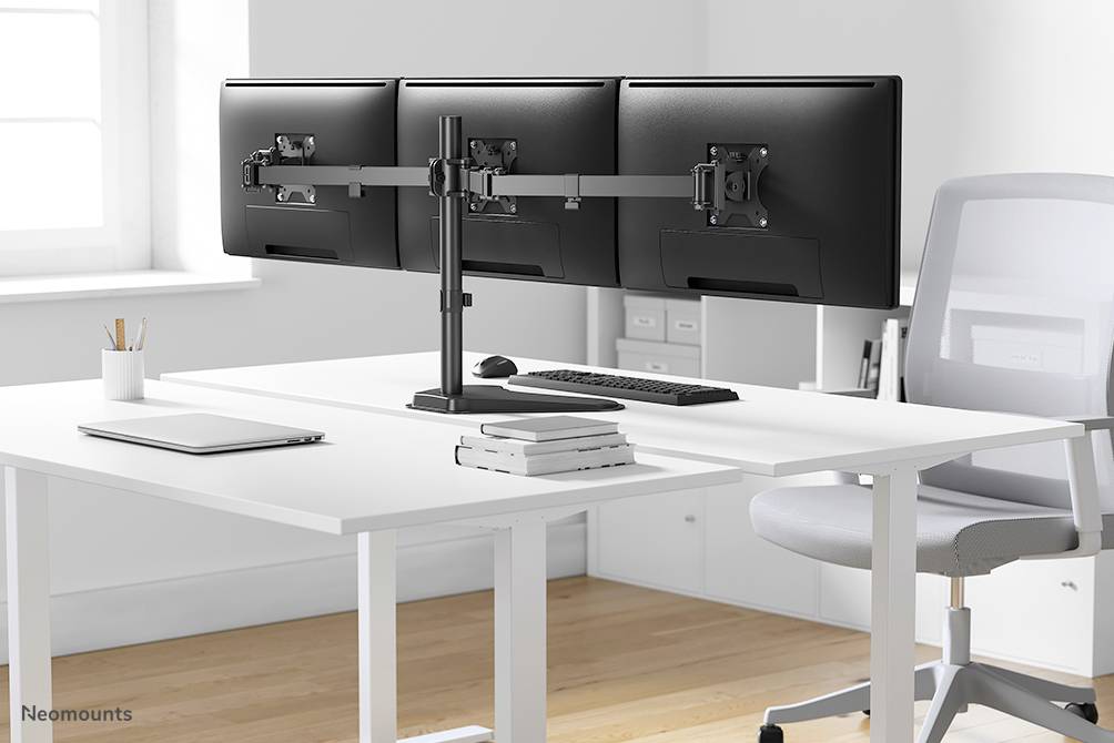 Three monitor screens on a mounting system on a modern white desk with keyboard, mouse and stack of books.