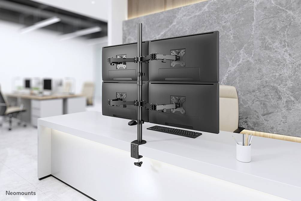 An office with a desk featuring a monitor mount holding four screens. In the background, additional workstations are visible.