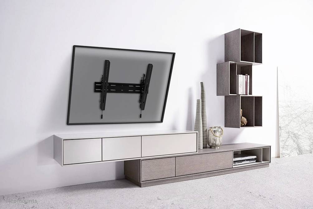 A living room with a light-coloured wall, where a tilted television is mounted on a bracket. Beside it, a modern shelving unit with decorative items.