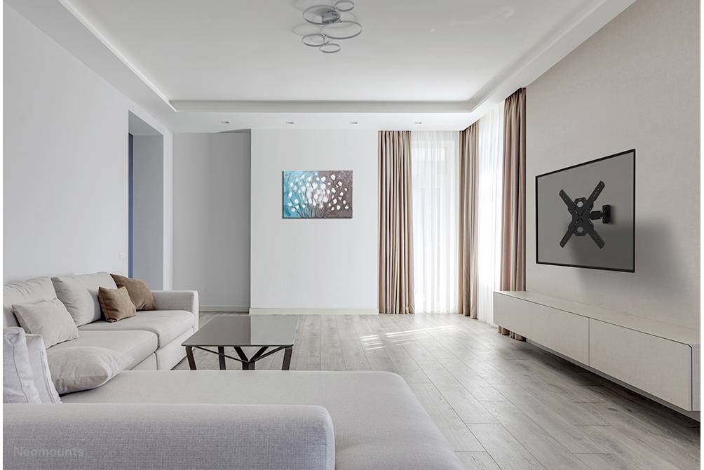 Bright living room with a grey sofa, coffee table, wall decor and TV mount on the wall. Large windows with beige curtains.