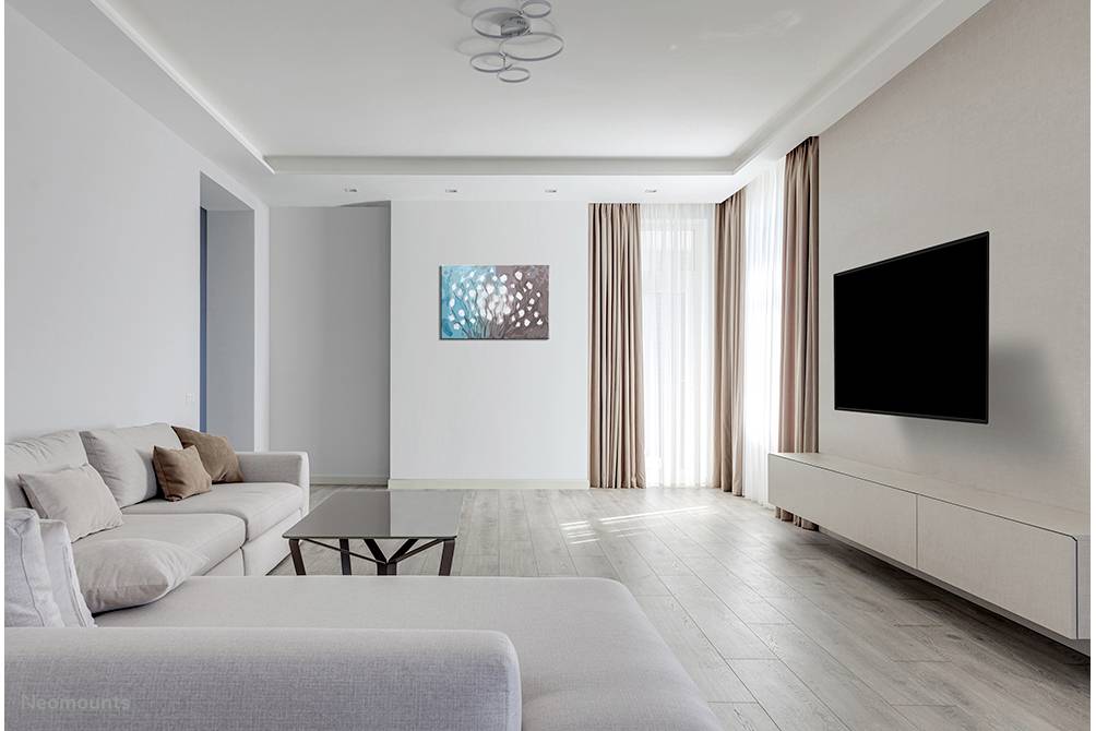 Bright living room with a grey sofa, large black flatscreen television mounted on the wall, wooden flooring and natural daylight streaming through large windows.