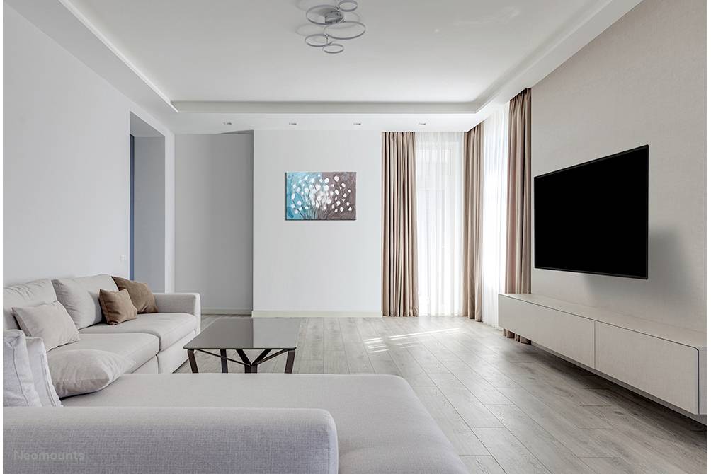 Bright living room with a beige sofa, coffee table, large television mounted on the wall and an abstract artwork. Large windows with curtains.