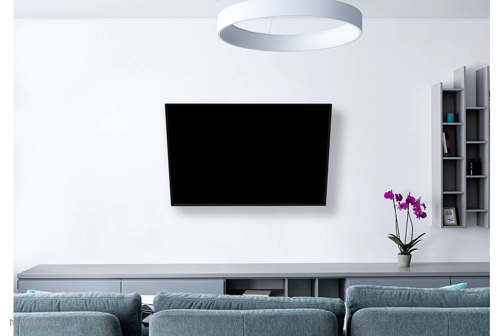 A modern living room with a large, black flatscreen television mounted on the white wall. In the foreground are blue sofas. To the right, a vase contains a purple orchid.