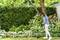 A woman trims garden edges with an electric trimmer next to lush green bushes and pink flowering plants on a sunny day.