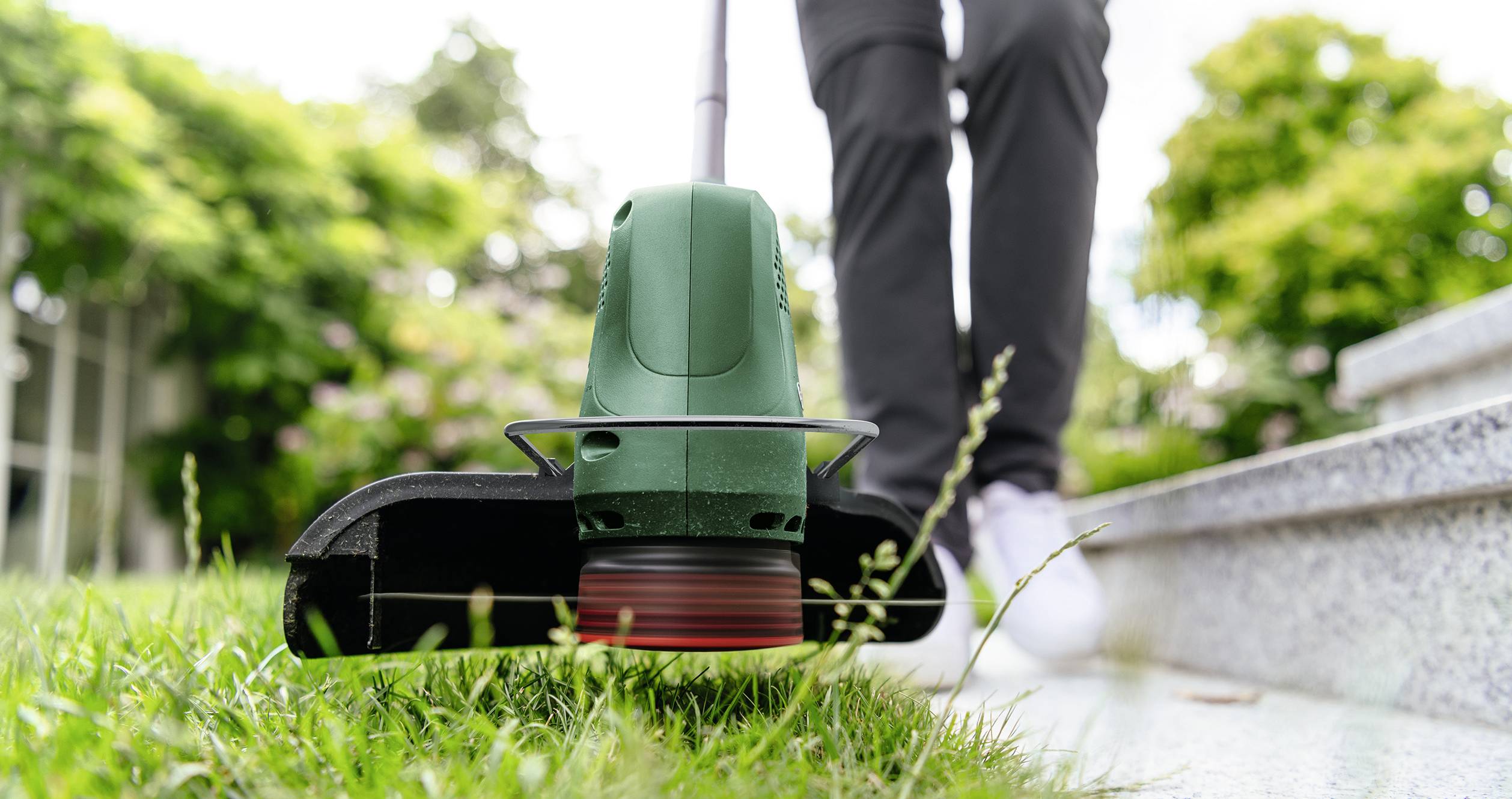 Person using a green electric grass trimmer on a lawn near steps, with greenery in the background.