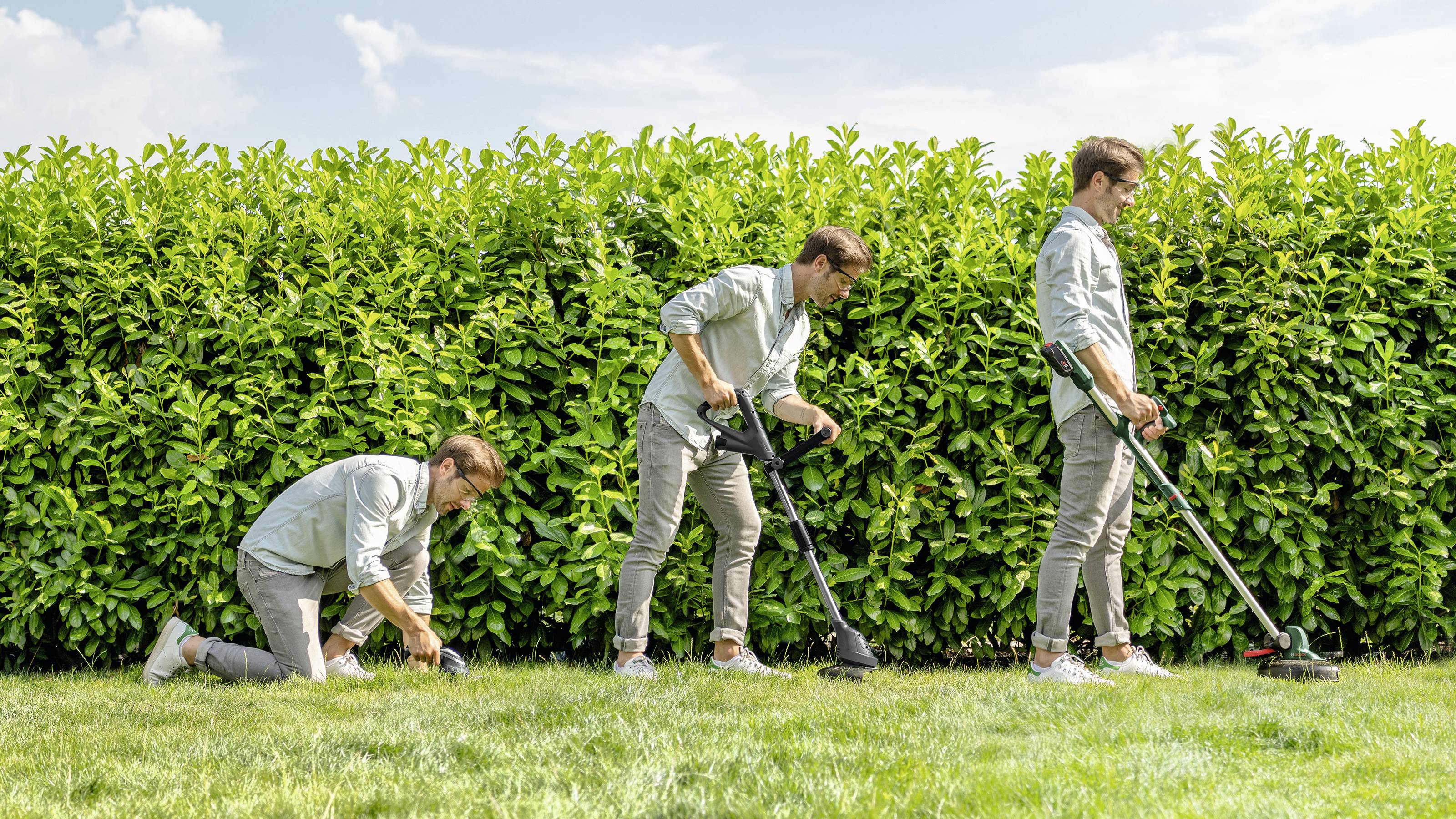 A man is mowing grass with a lawn trimmer in front of a tall hedge. He is wearing a light shirt, grey trousers, and sunglasses.