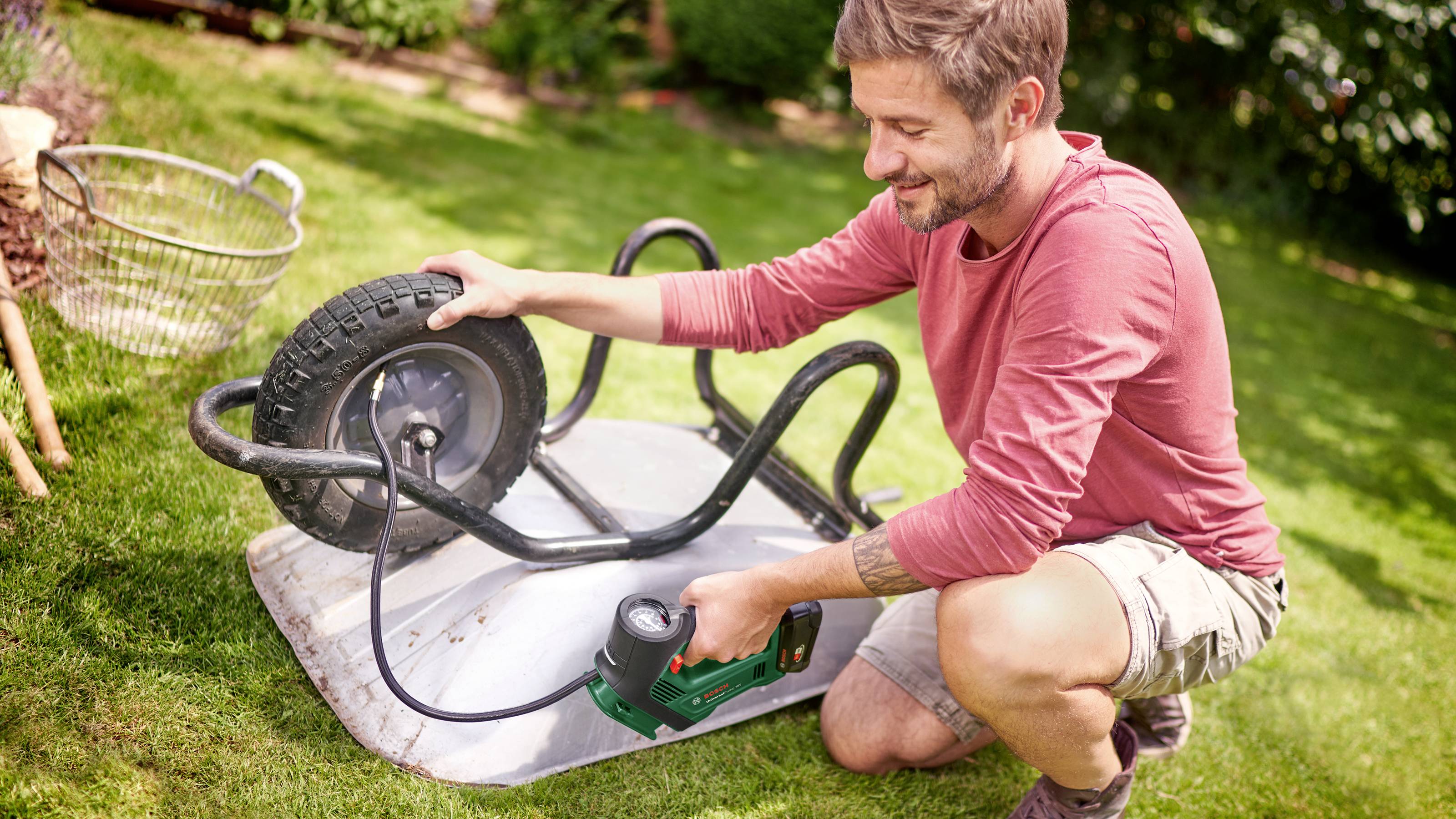 A man is kneeling in the garden and pumping the tyre of an upturned wheelbarrow. Gardening tools can be seen in the background.