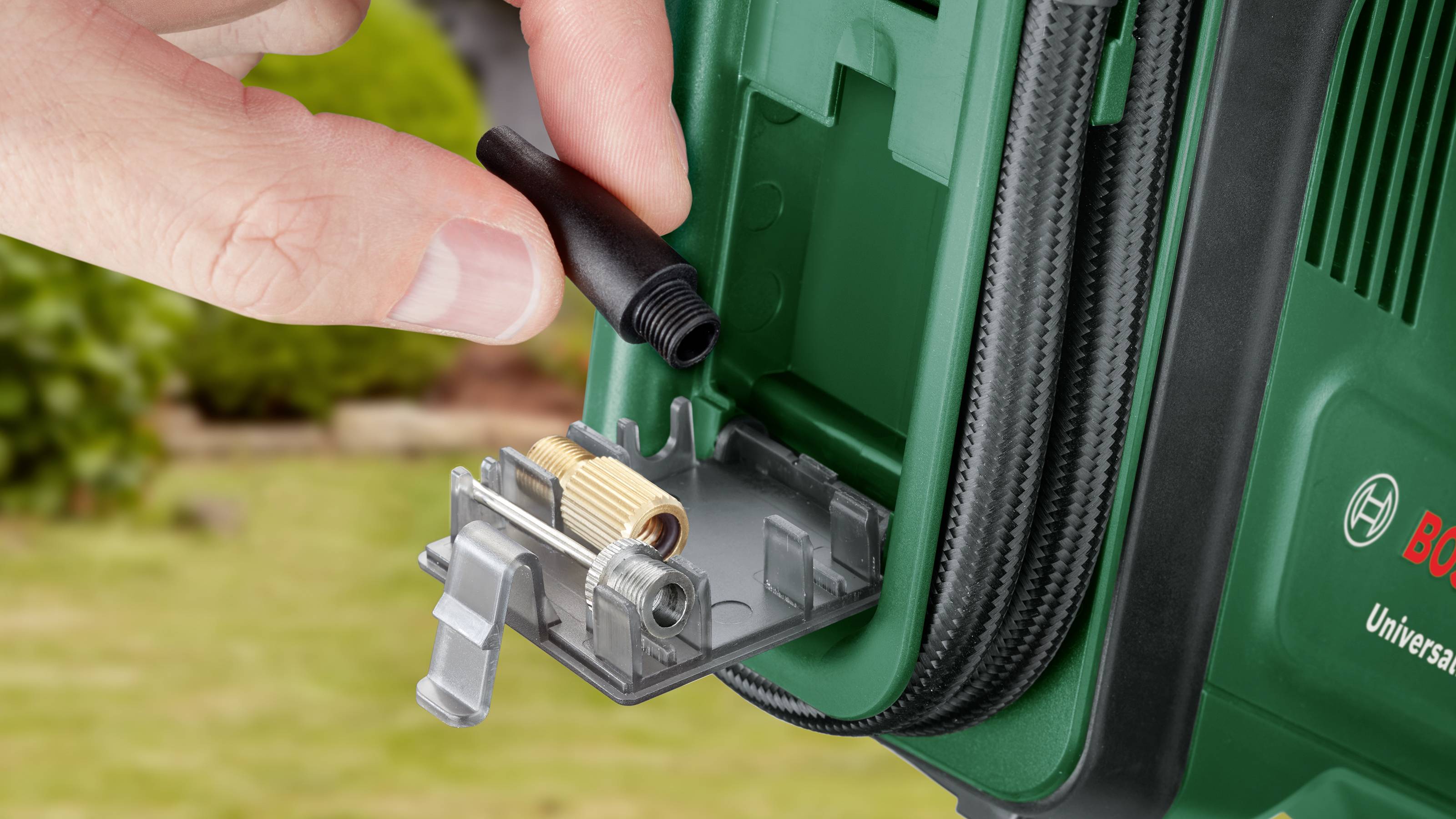 A person is holding a small accessory part next to an open compartment of a garden tool. Various attachments are located in the compartment.