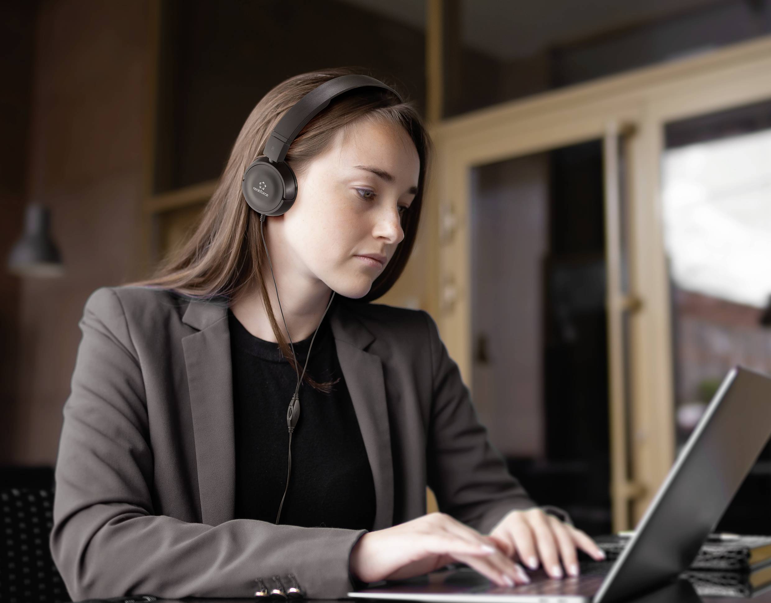 A woman in business attire is working intently on a laptop while wearing headphones. She is seated in a modern office.