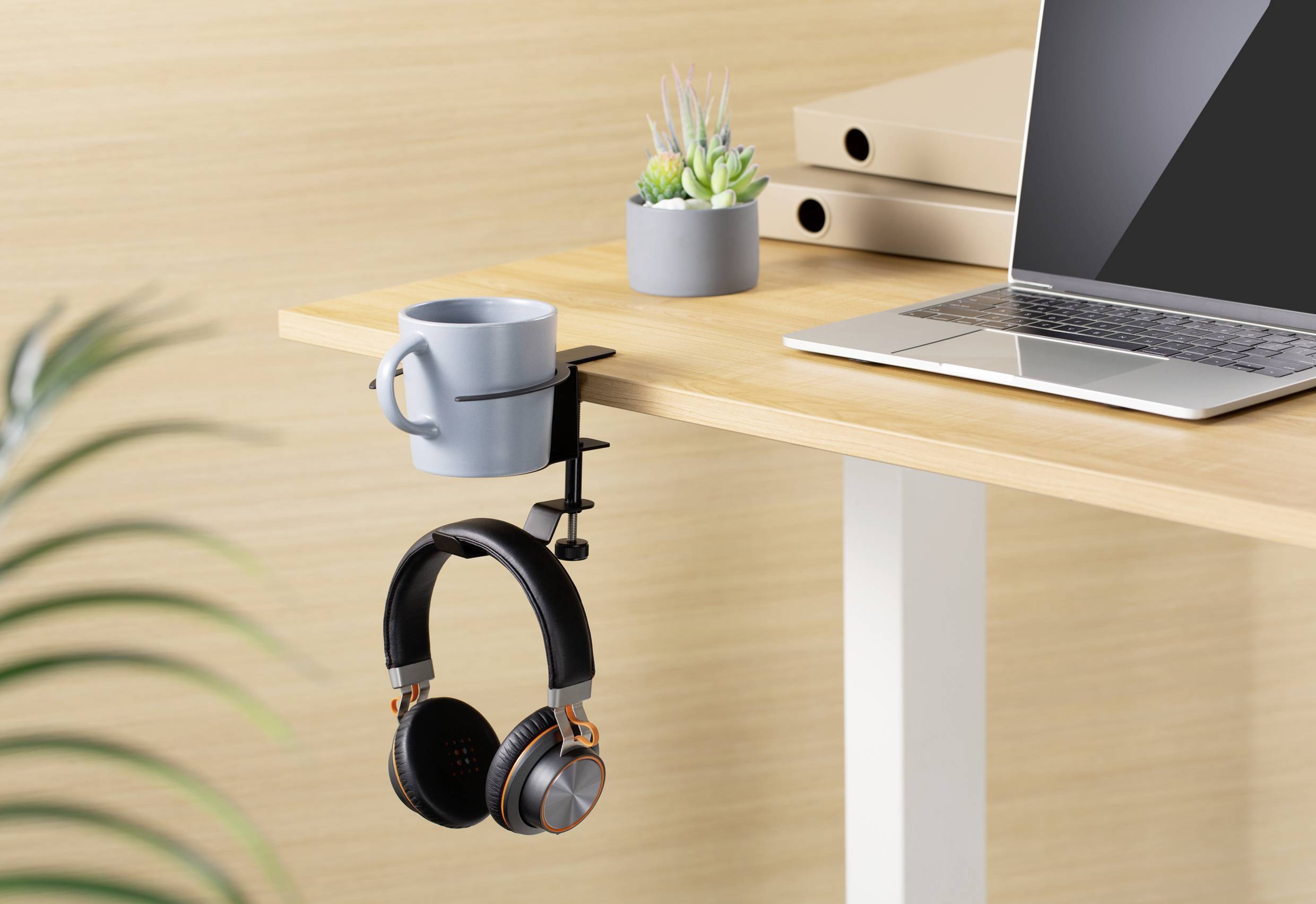 A table with a laptop, a cactus, and a headphone stand holding a mug and headphones at the table's edge.