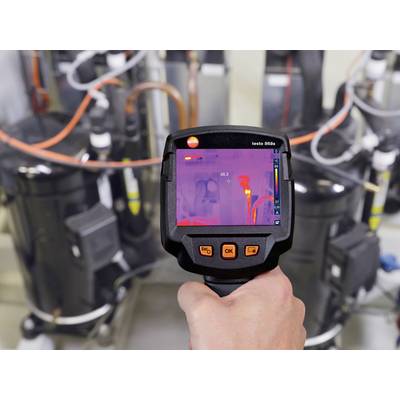 A person is holding a thermal imaging camera displaying a temperature measurement. The background shows technical equipment.