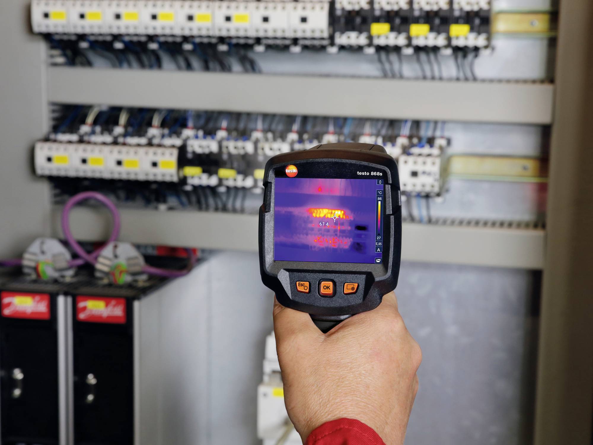 A person is holding a thermal imaging camera in front of an open electrical cabinet. The screen displays temperature variations across the components.