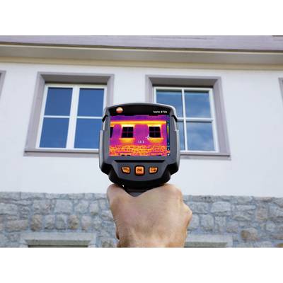 A hand is holding a thermal imaging camera, showing a building with two windows. Temperature variations are visible on the display.