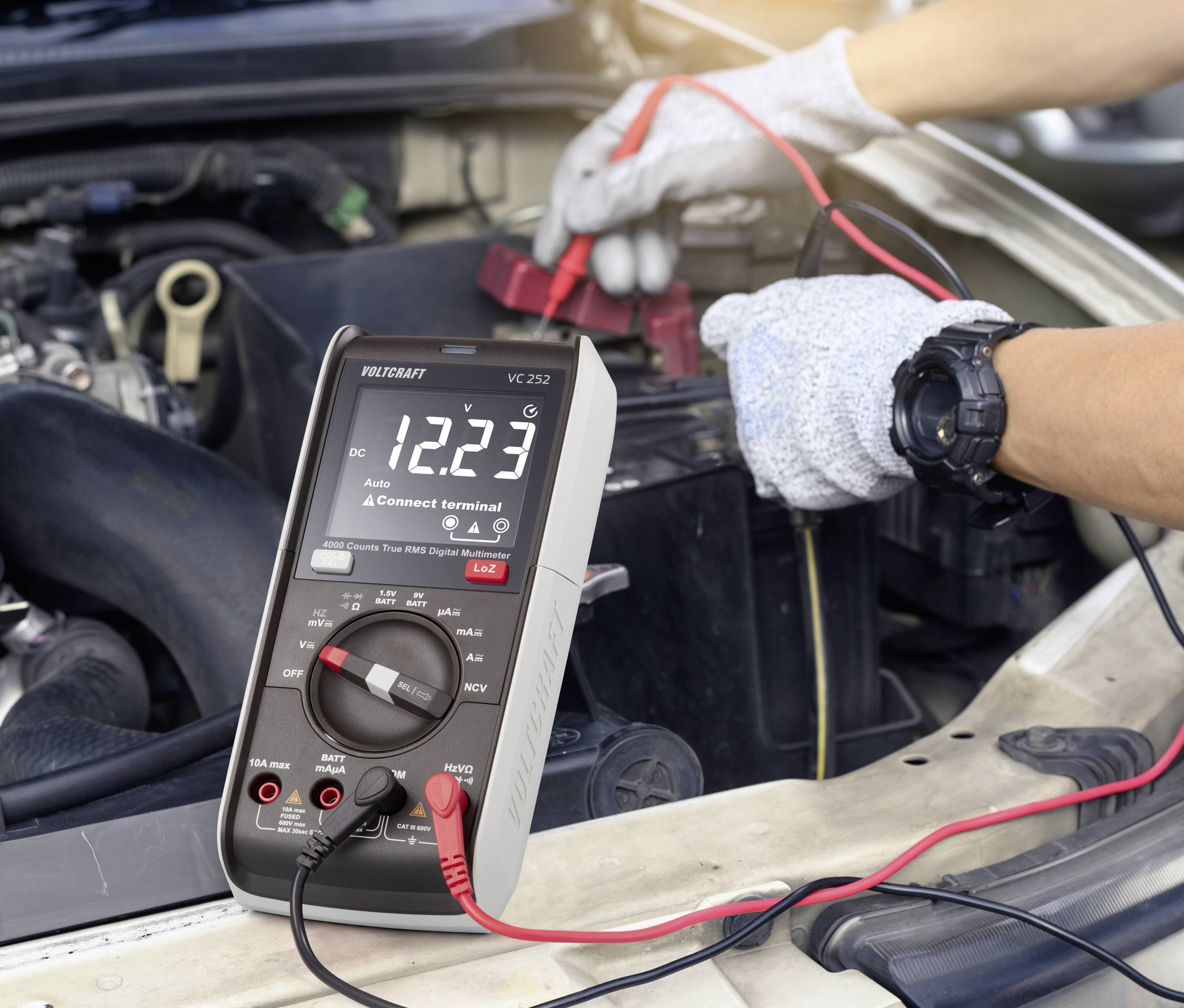 A multimeter is showing 12.23 volts while connected to a car battery in the engine compartment. A person wearing gloves is holding the clamps.