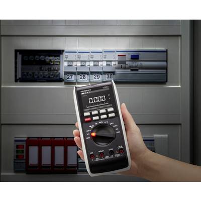 'A hand holds a multimeter, displaying 0.00, in front of an opened electrical distribution board with switches and fuses, symbolising electrical testing.'