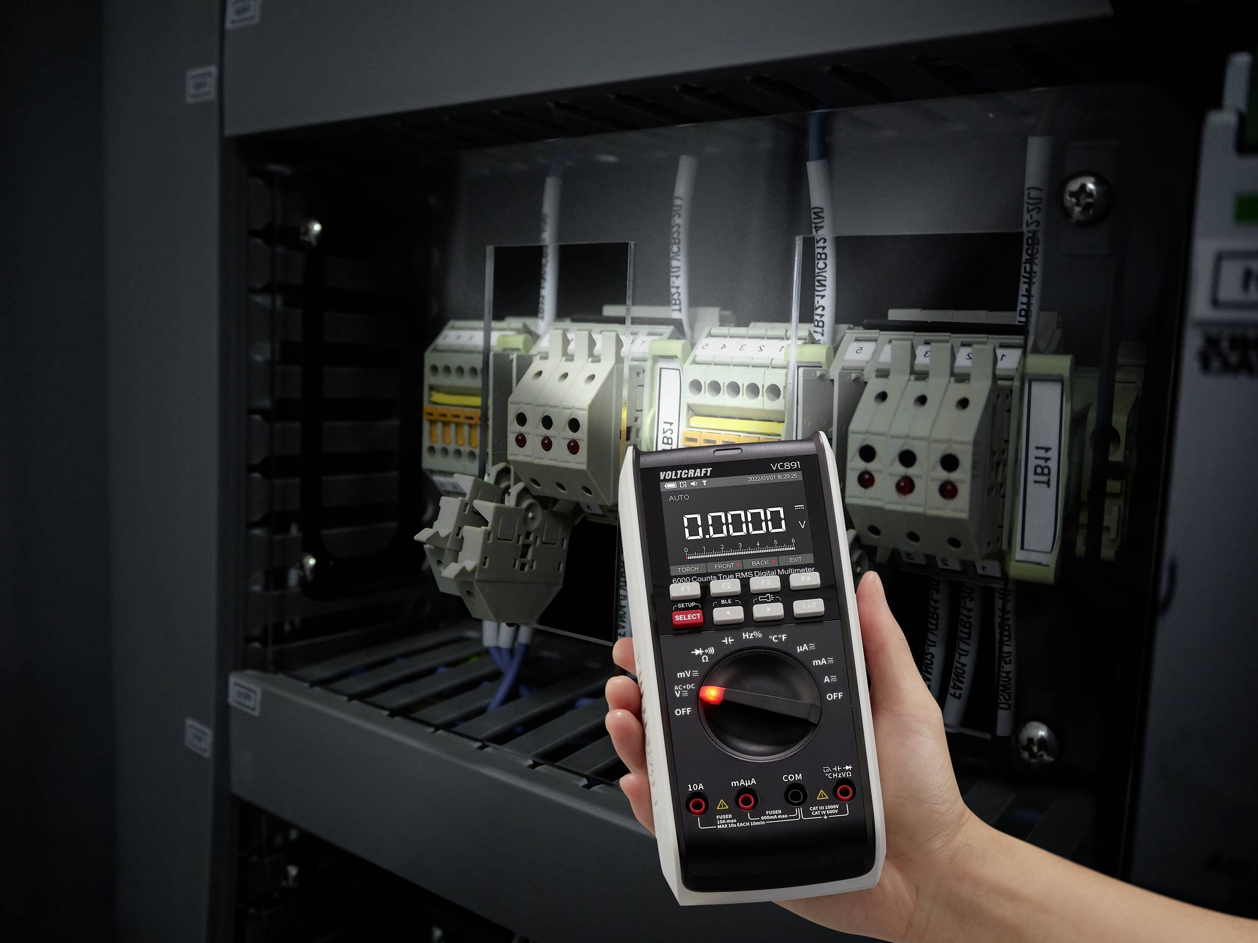 A hand is holding a multimeter in front of an electrical cabinet, with several electrical components and cables visible.