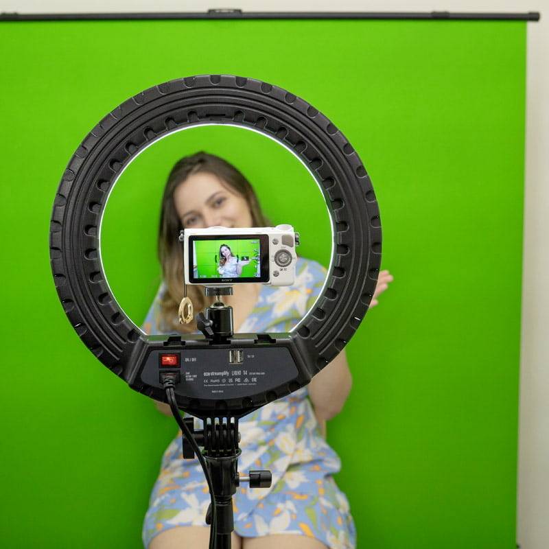 A person is sitting in front of a green background, filmed by a camera mounted on a ring light.