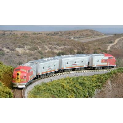 A model train in red and silver paintwork travels along a curved track through hilly, vegetation-rich countryside.