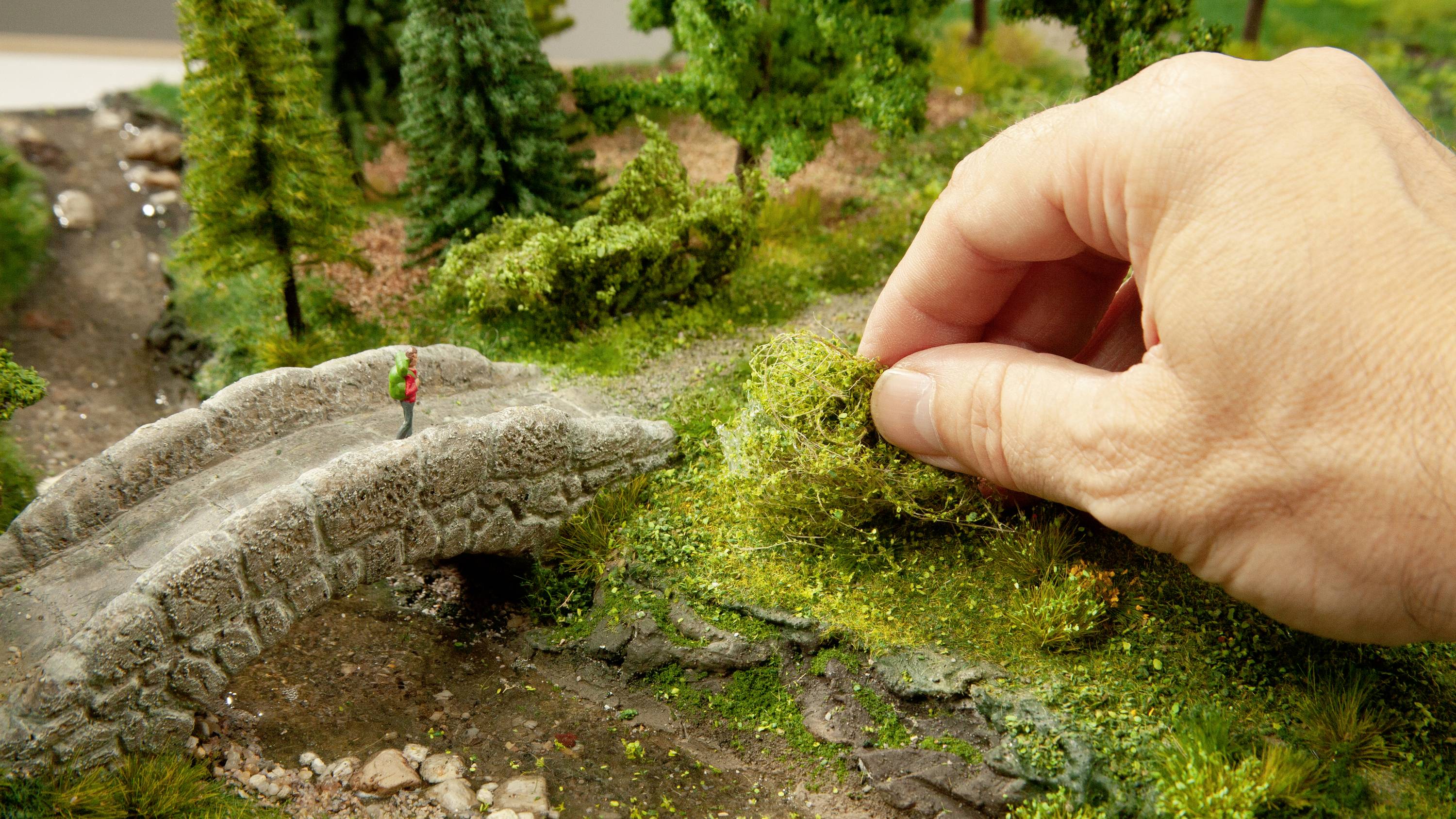 A hand places botanical decoration on a detailed miniature model featuring a small stone bridge and a wanderer.