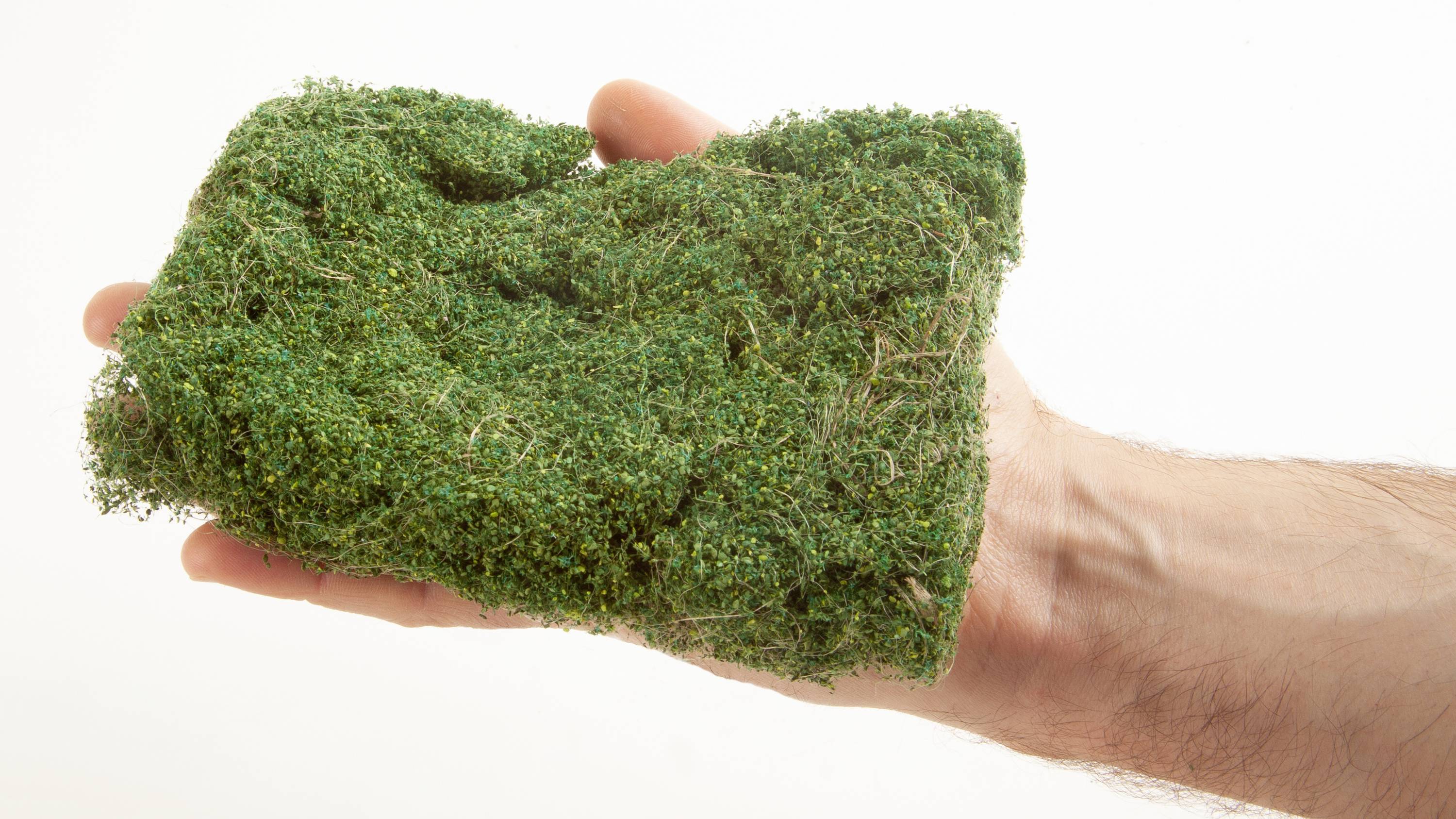 A hand holds a rectangular piece of green moss that looks artificial. It is presented against a white background.