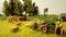 Farmers harvesting hay in a field with tractors and hay bales, surrounded by trees. A rural scene with agricultural machinery.