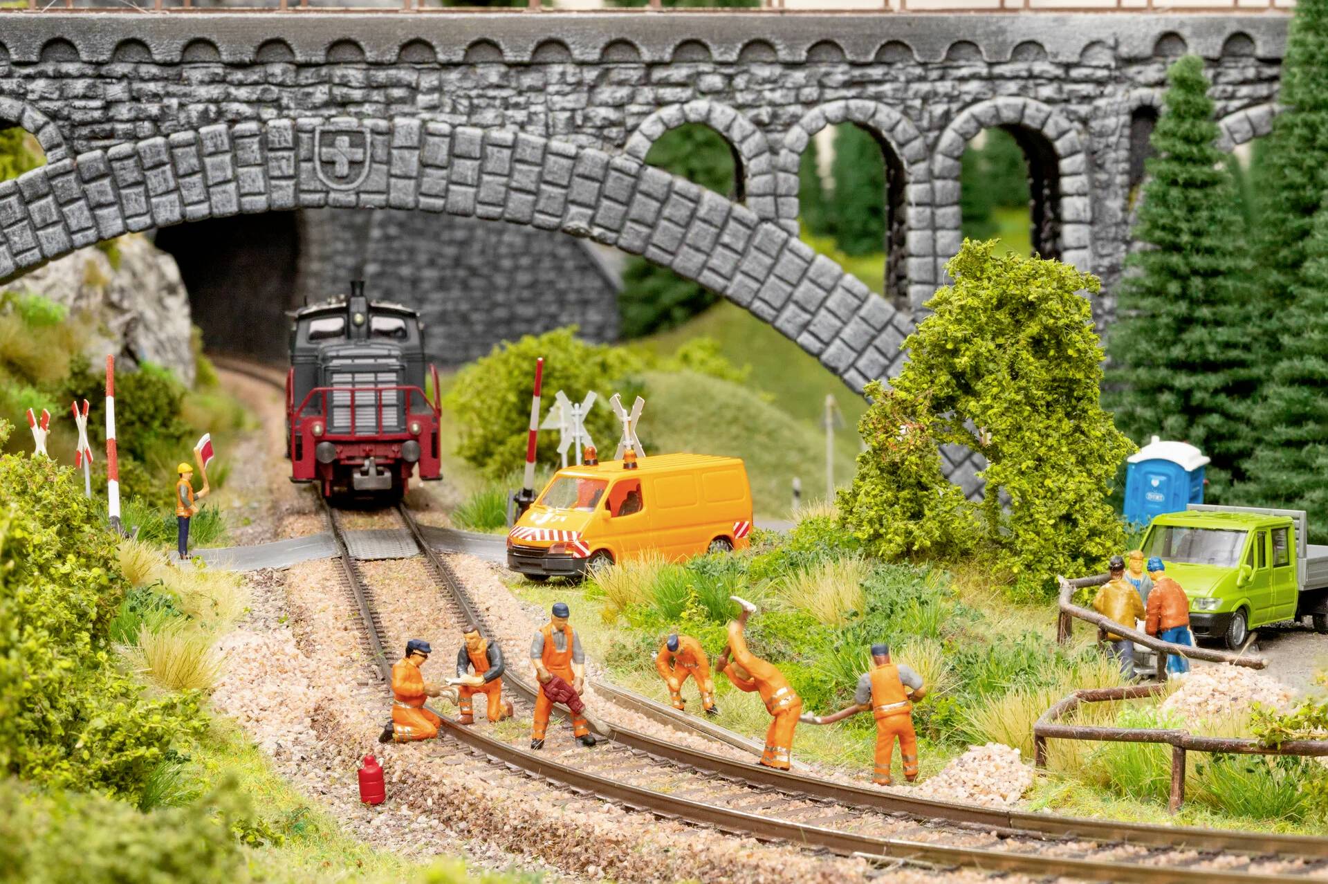 Miniature railway scene with workers in orange clothing on the tracks, surrounded by trees, a yellow van and a stone bridge.