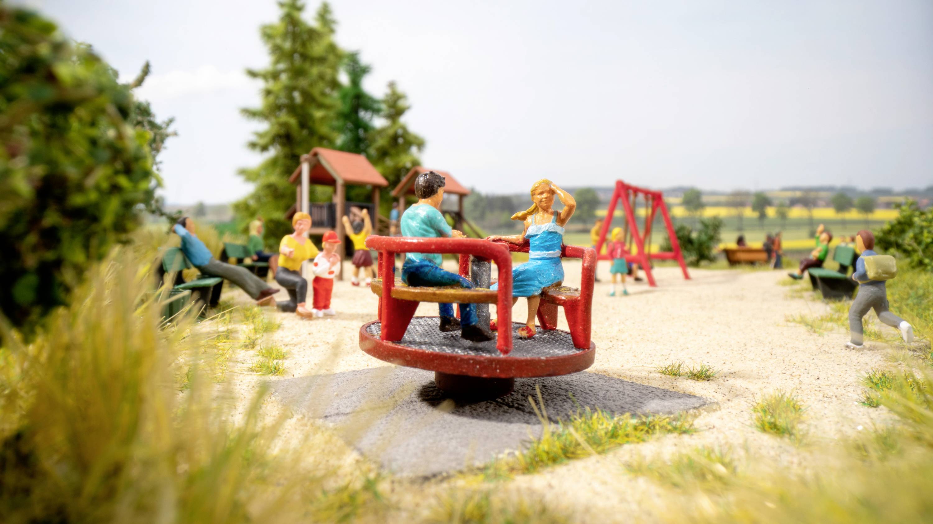 Children are playing on a roundabout in a park with green trees and swings in the background. A sunny day.