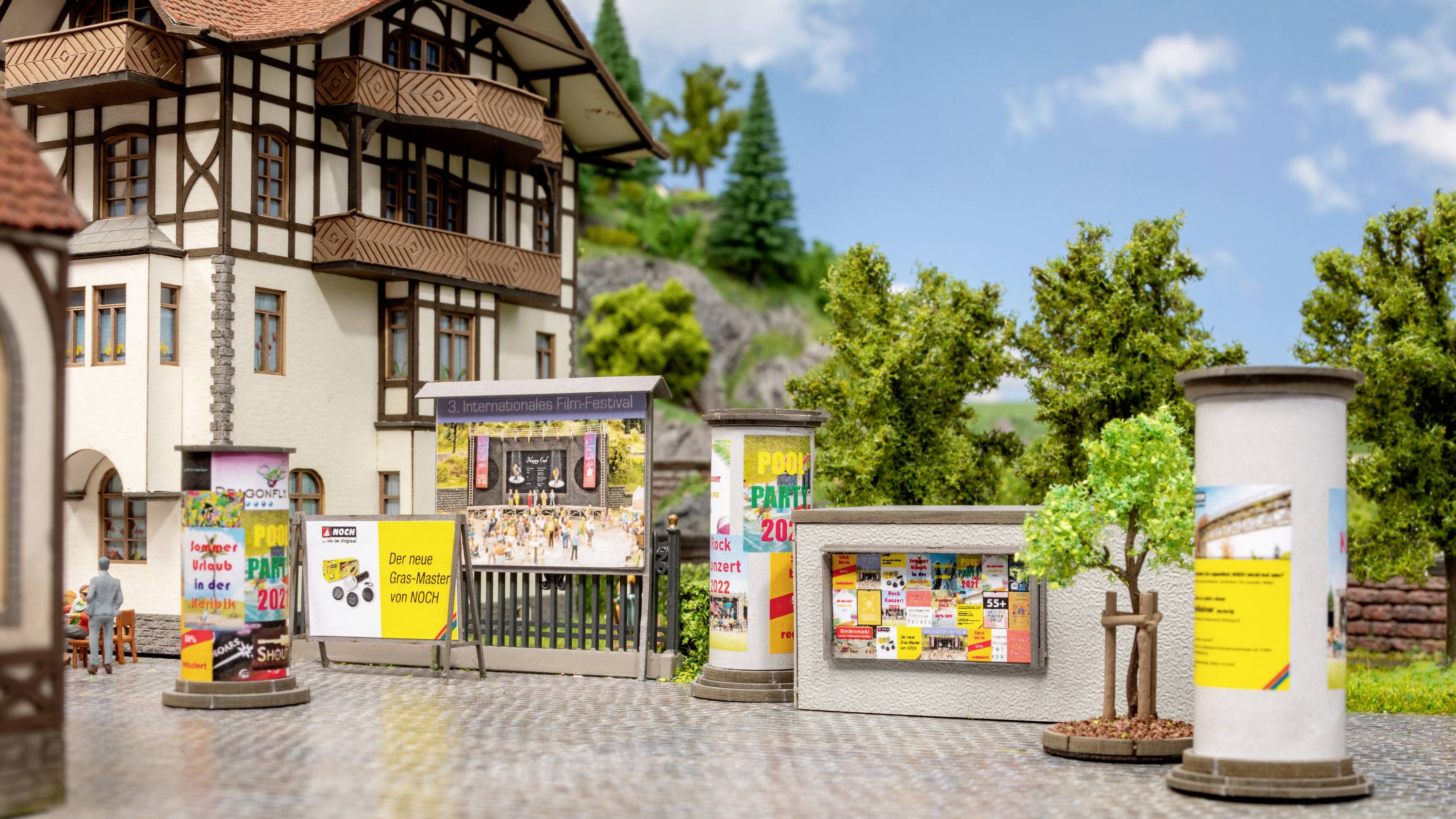 A model street scene with billboard walls and advertising boards in front of a half-timbered house, surrounded by trees and hills.